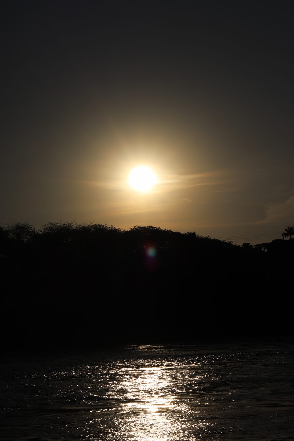 A golden sun hangs low in a hazy sky, casting a bright, glowing reflection across the Source of the Nile
