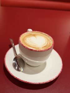 White cappuccino cup with heart-shaped latte art on a matching saucer with a silver spoon, set on a red table.