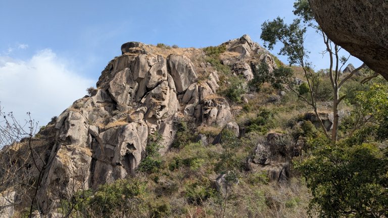 Rugged hillside covered with large gray boulders and rocks, with sparse green shrubs and trees under a bright blue sky with scattered clouds.