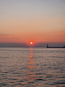 Vertical photograph of a bright orange sun setting low over calm ocean waters with a subtle lighthouse silhouette on the right.
