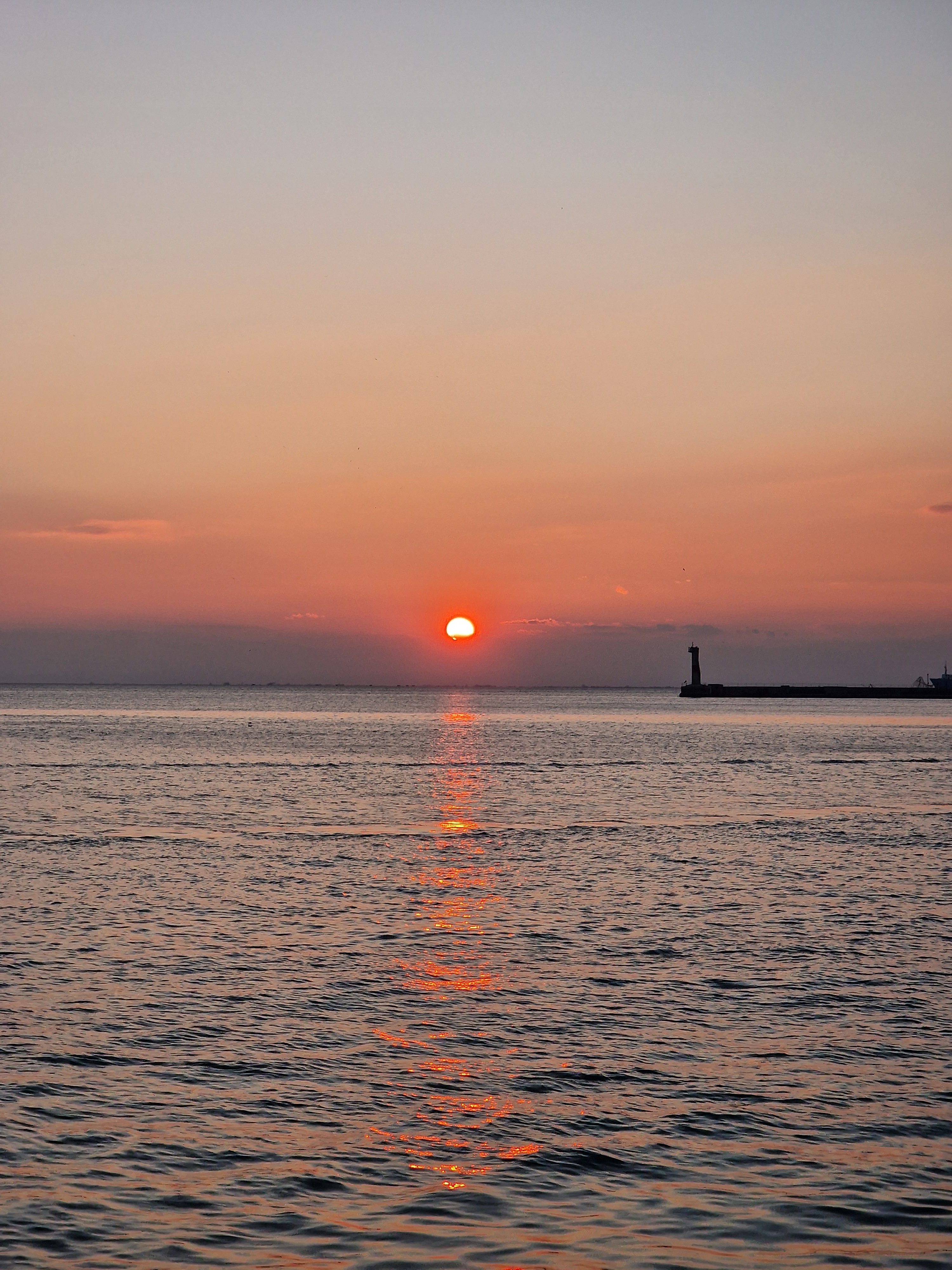 Vertical photograph of a bright orange sun setting low over calm ocean waters with a subtle lighthouse silhouette on the right.