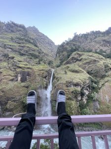 A POV shot looking down at legs wearing black trousers, socks, and slides, resting on a pink metal railing over a gushing waterfall plunging down a steep, grassy mountain gorge.