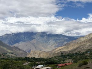 A wide shot of a green mountain valley with terraced fields and cluster settlements, framed by massive arid, cloud-draped mountain ridges.