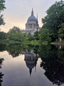 Symmetrical water reflection of St Paul's Cathedral surrounded by green trees during sunset in London.