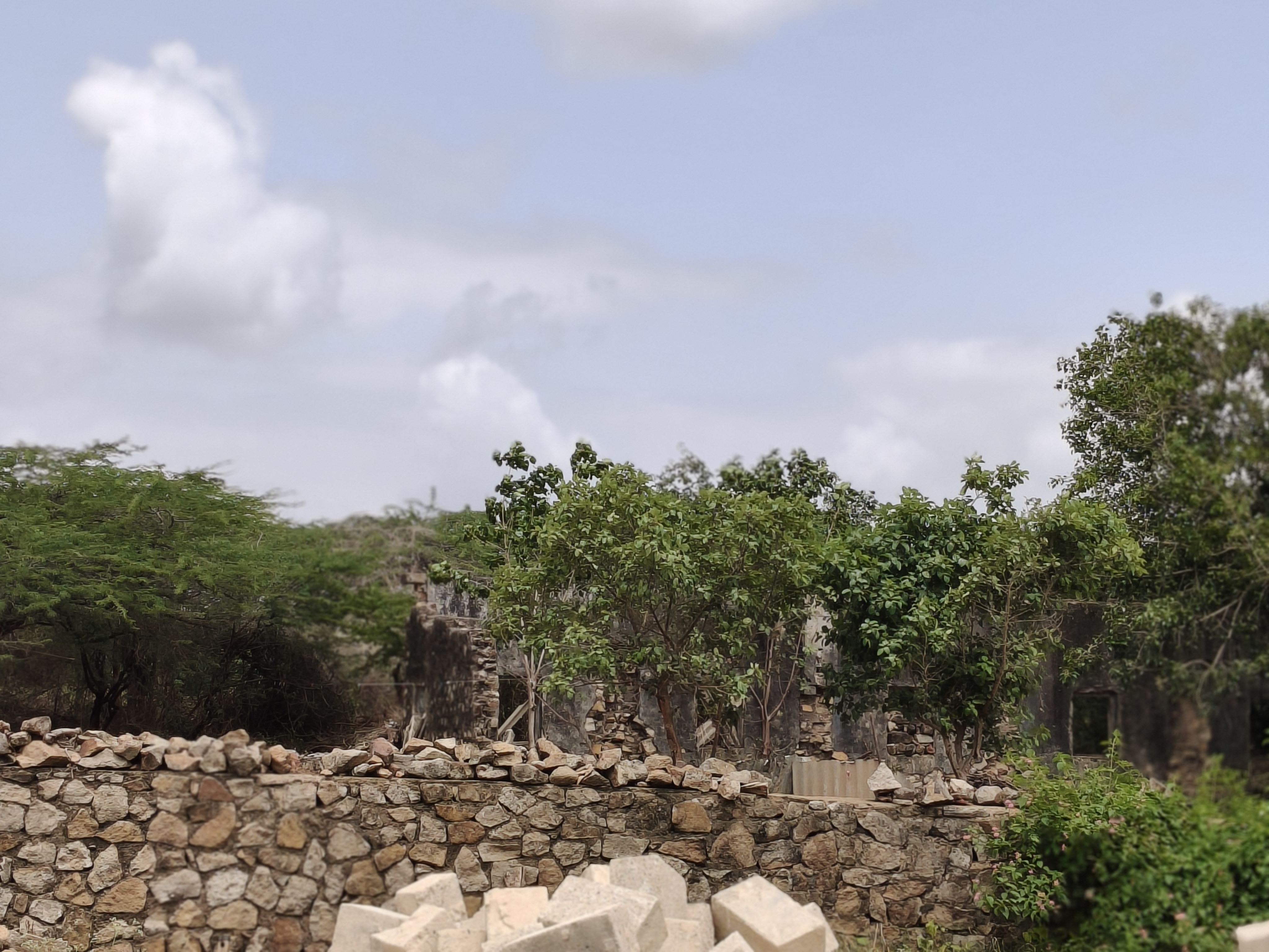 Low stone wall with green trees and a ruined building in the background under a partly cloudy sky.