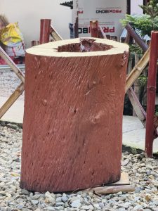 Large hollow wooden log resting on small pebbles outdoors, with bamboo poles and greenery in the background.