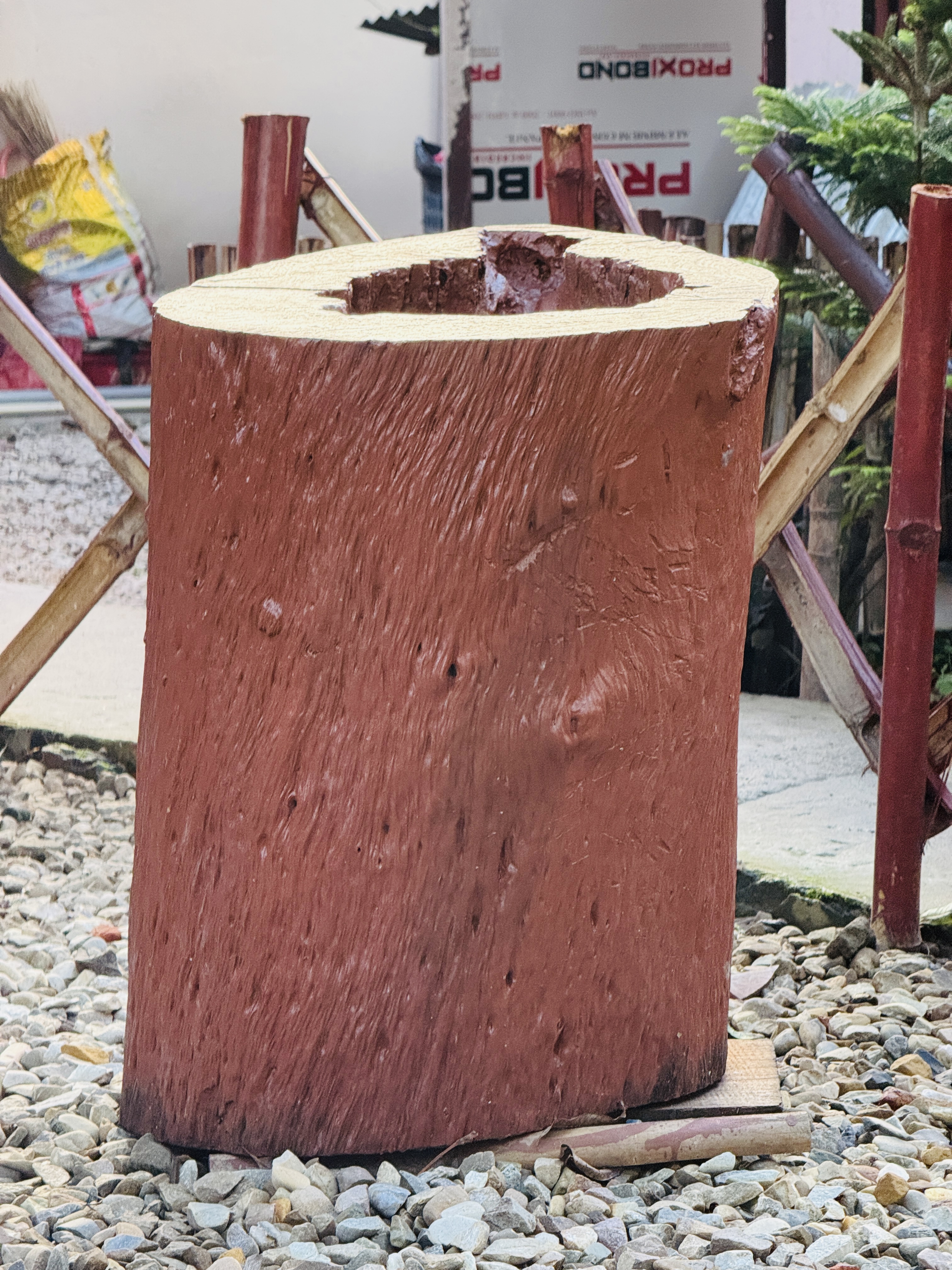 Large hollow wooden log resting on small pebbles outdoors, with bamboo poles and greenery in the background.