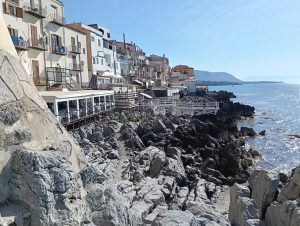 A coastal scene showcasing a rocky shoreline with various sizes of dark, jagged rocks jutting out into the calm sea. The sun illuminates the scene, creating a bright and inviting atmosphere, while the gentle waves of the sea reflect the blue sky. In the distance, faint hills can be seen, adding depth to the picturesque view.