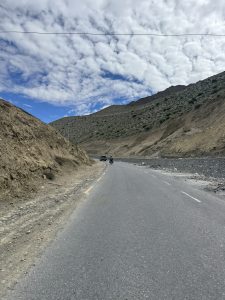 Curving mountain road through a dry canyon with a motorcycle and SUV under a cloudy blue sky.