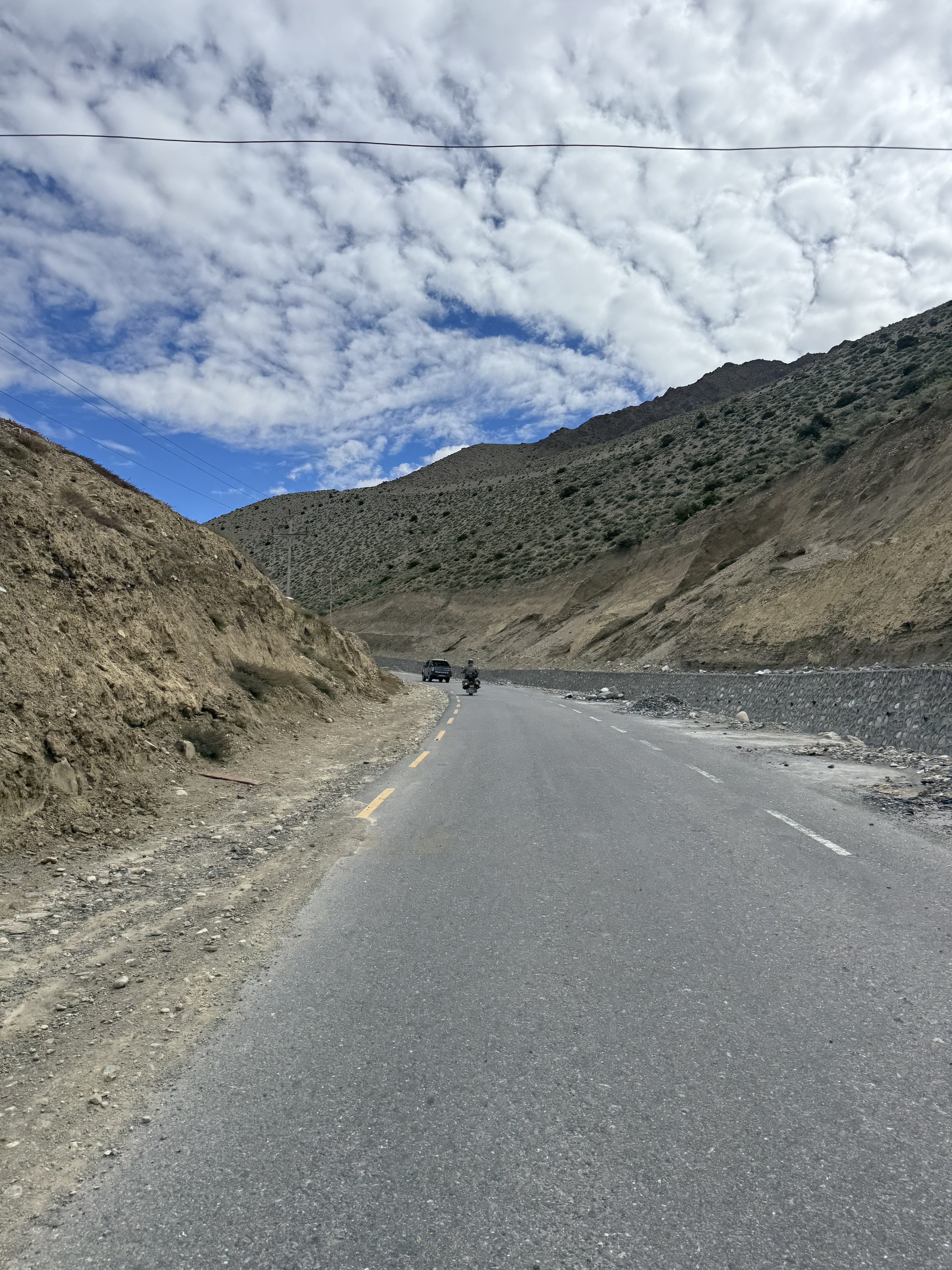Curving mountain road through a dry canyon with a motorcycle and SUV under a cloudy blue sky.