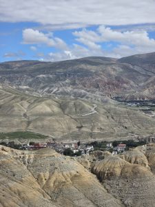 Elevated view of dry, eroded mountains and a small settlement under a partly cloudy sky.