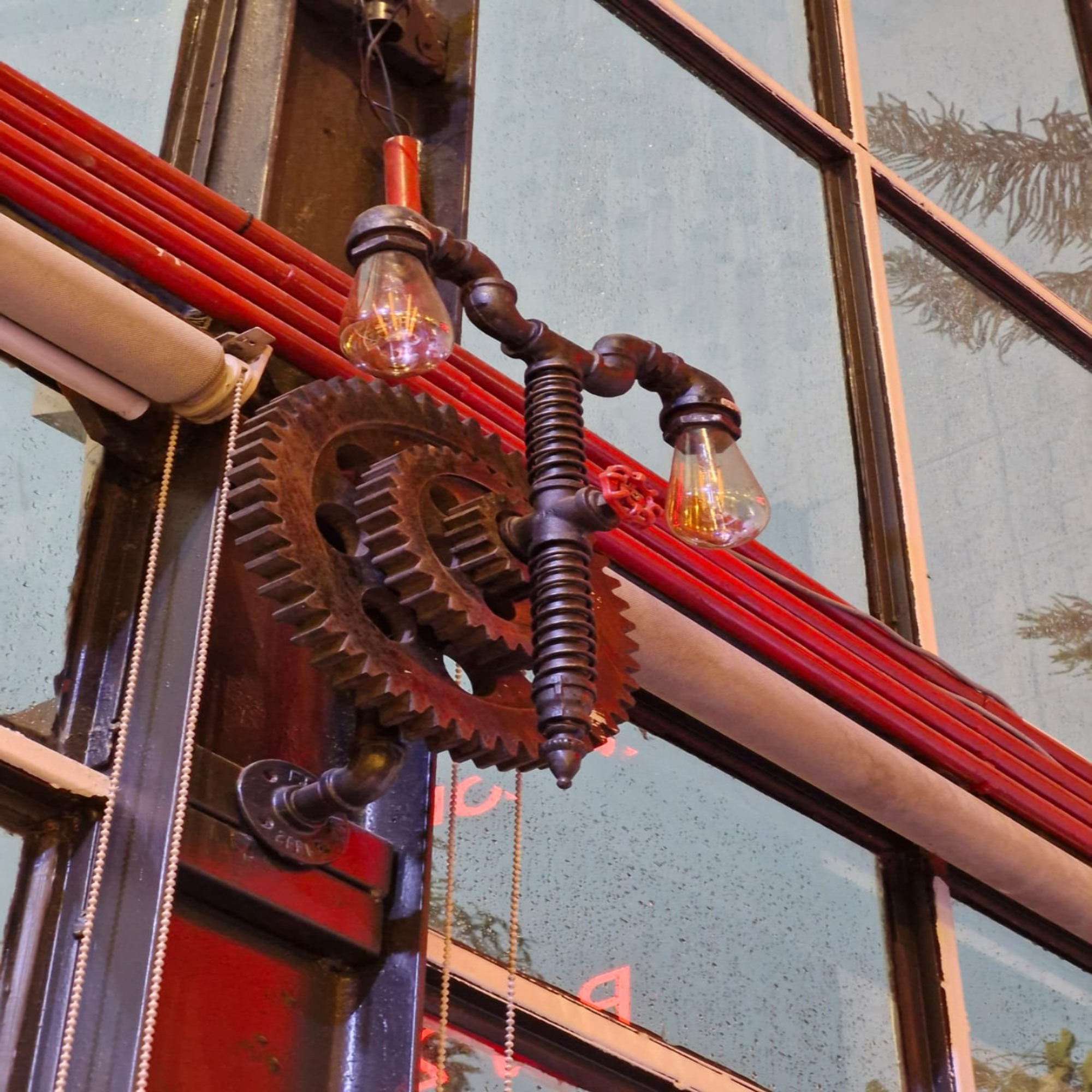 Vintage industrial wall fixture with interlocking gears, red pipes, and glowing Edison bulbs against a weathered window.