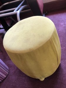 A small, round yellow cushioned stool with a soft, padded seat and cylindrical shape, placed indoors on a purple carpet.
