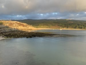 Panoramic view of the bay along the Camiño dos Faros in Corme, with A Arnela Beach on the left and golden sunset light over A Hermida Beach, the surrounding hills, and boats on calm water.
