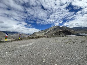 A wide, pebbled ground stretches into the distance, lined with colorful vertical prayer flags. In the background, stark, arid mountain ranges rise beneath a vivid blue sky filled with textured, broken white clouds.