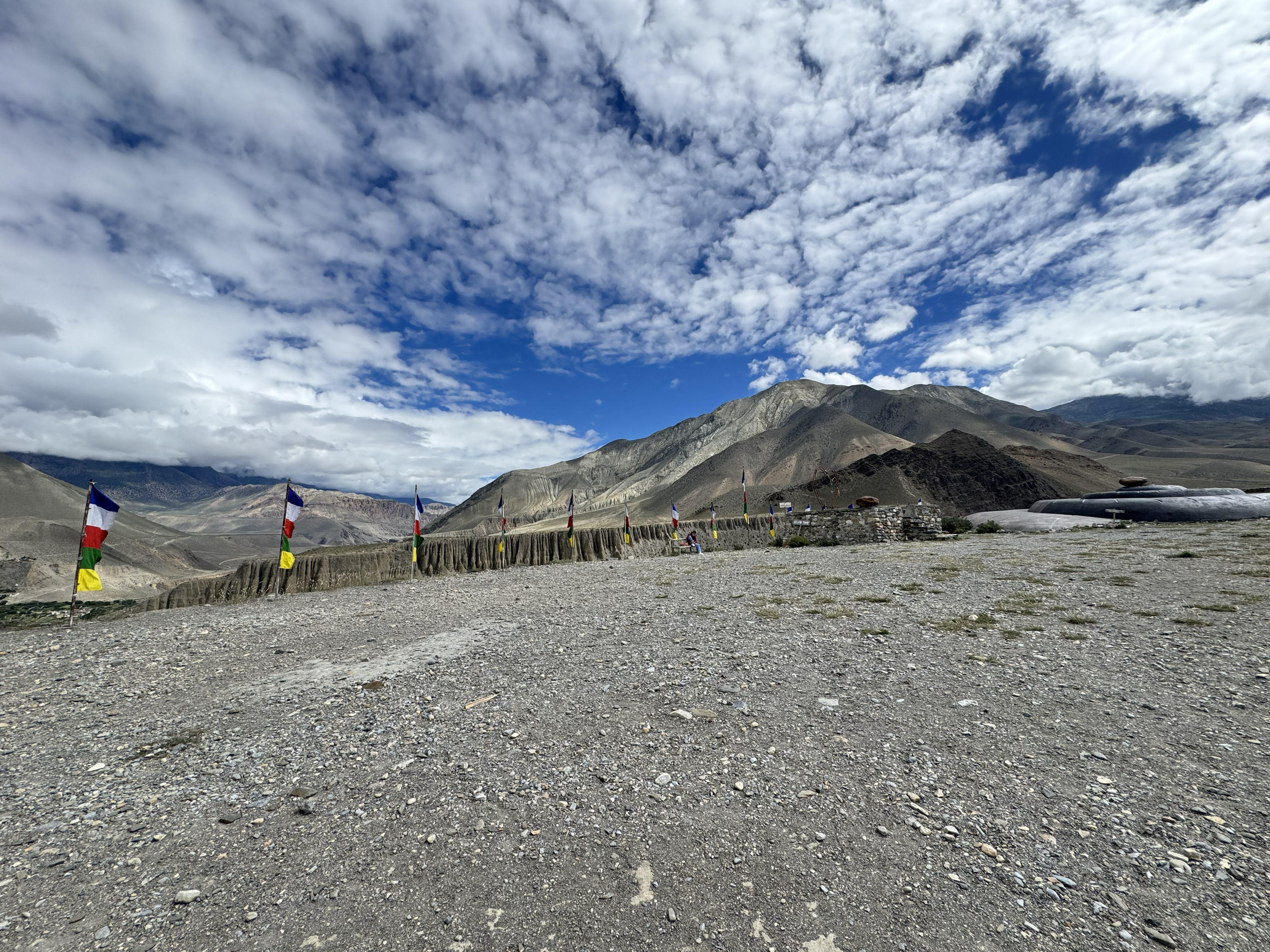 A wide, pebbled ground stretches into the distance, lined with colorful vertical prayer flags. In the background, stark, arid mountain ranges rise beneath a vivid blue sky filled with textured, broken white clouds.