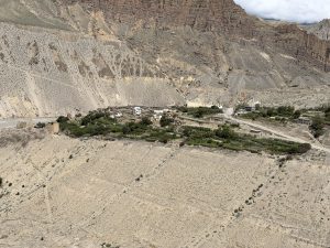 A high-angle view of a secluded mountain village with terraced greenery nestled along the edge of a dry, light-colored gorge cliff.