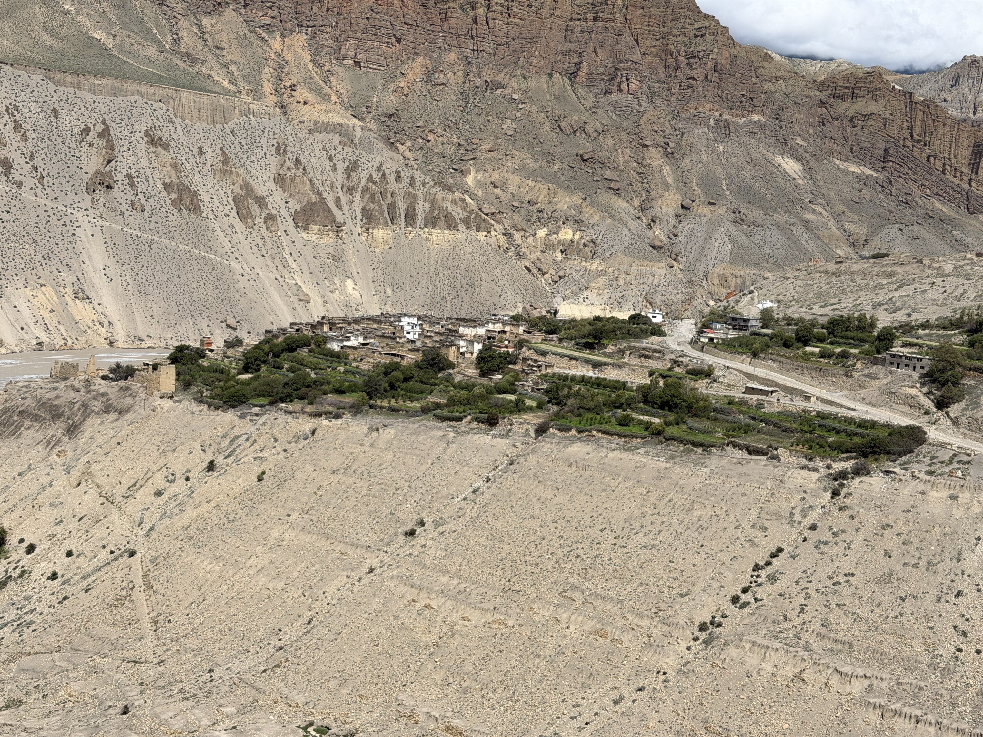 A high-angle view of a secluded mountain village with terraced greenery nestled along the edge of a dry, light-colored gorge cliff.
