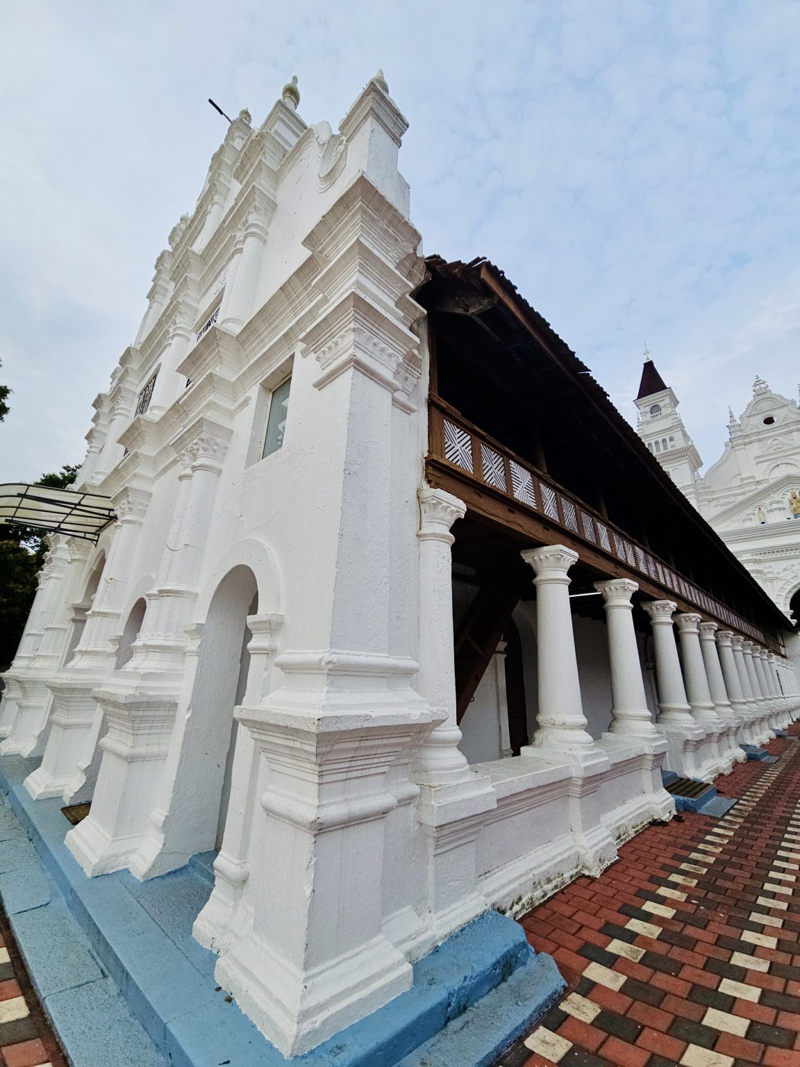 A side view of the church showing its ornate white façade, traditional wooden upper structure, rows of columns, and patterned walkway. The perspective effectively highlights the architecture. Location: Ramapuram, Kottayam, Kerala.