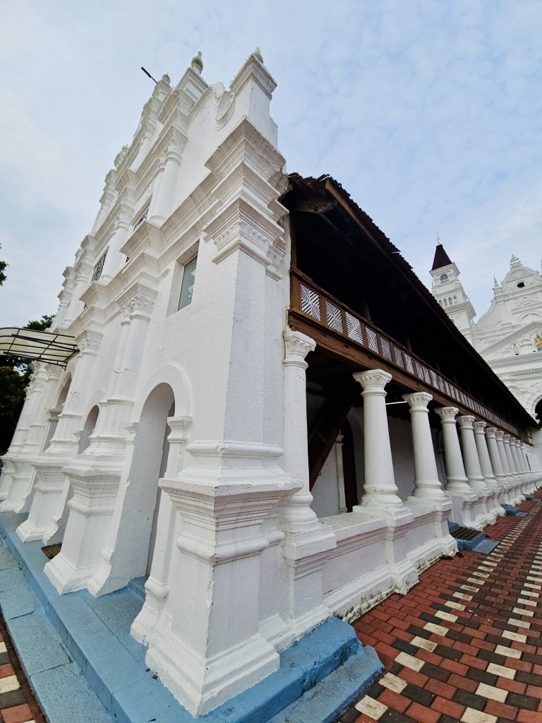 A side view of the church showing its ornate white façade, traditional wooden upper structure, rows of columns, and patterned walkway. The perspective effectively highlights the architecture. Location: Ramapuram, Kottayam, Kerala.