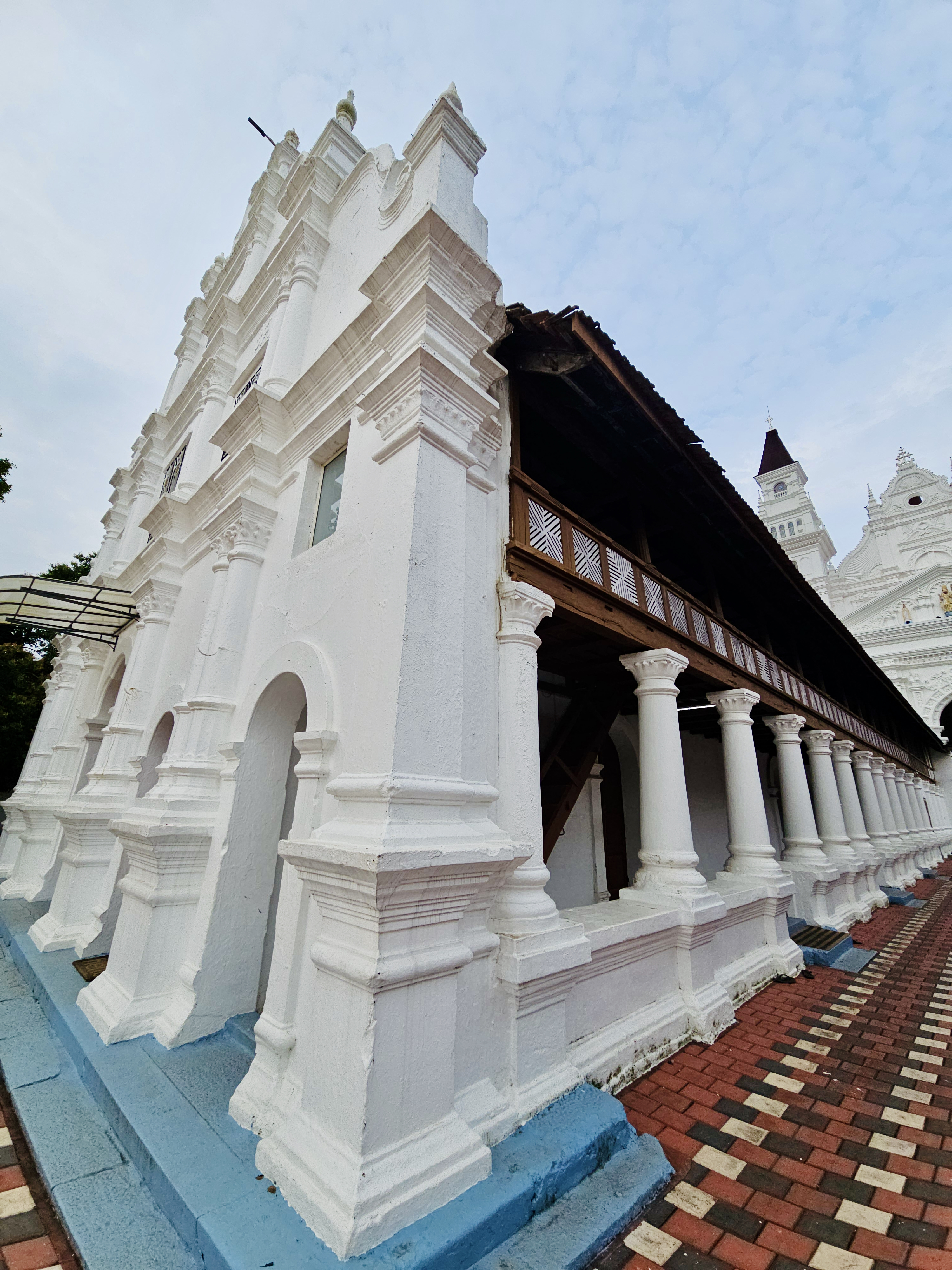 A side view of the church showing its ornate white façade, traditional wooden upper structure, rows of columns, and patterned walkway. The perspective effectively highlights the architecture. Location: Ramapuram, Kottayam, Kerala.