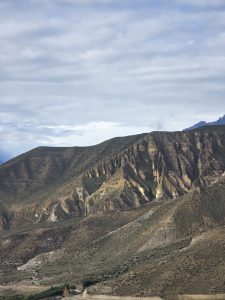 A wide landscape view shows steep, dry, eroded brown mountain slopes descending toward a valley floor on the right, under an overcast sky with light, hazy clouds.