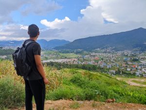 Person with a backpack standing on a hill overlooking a green valley, cityscape, and distant mountains under a partly cloudy sky.
