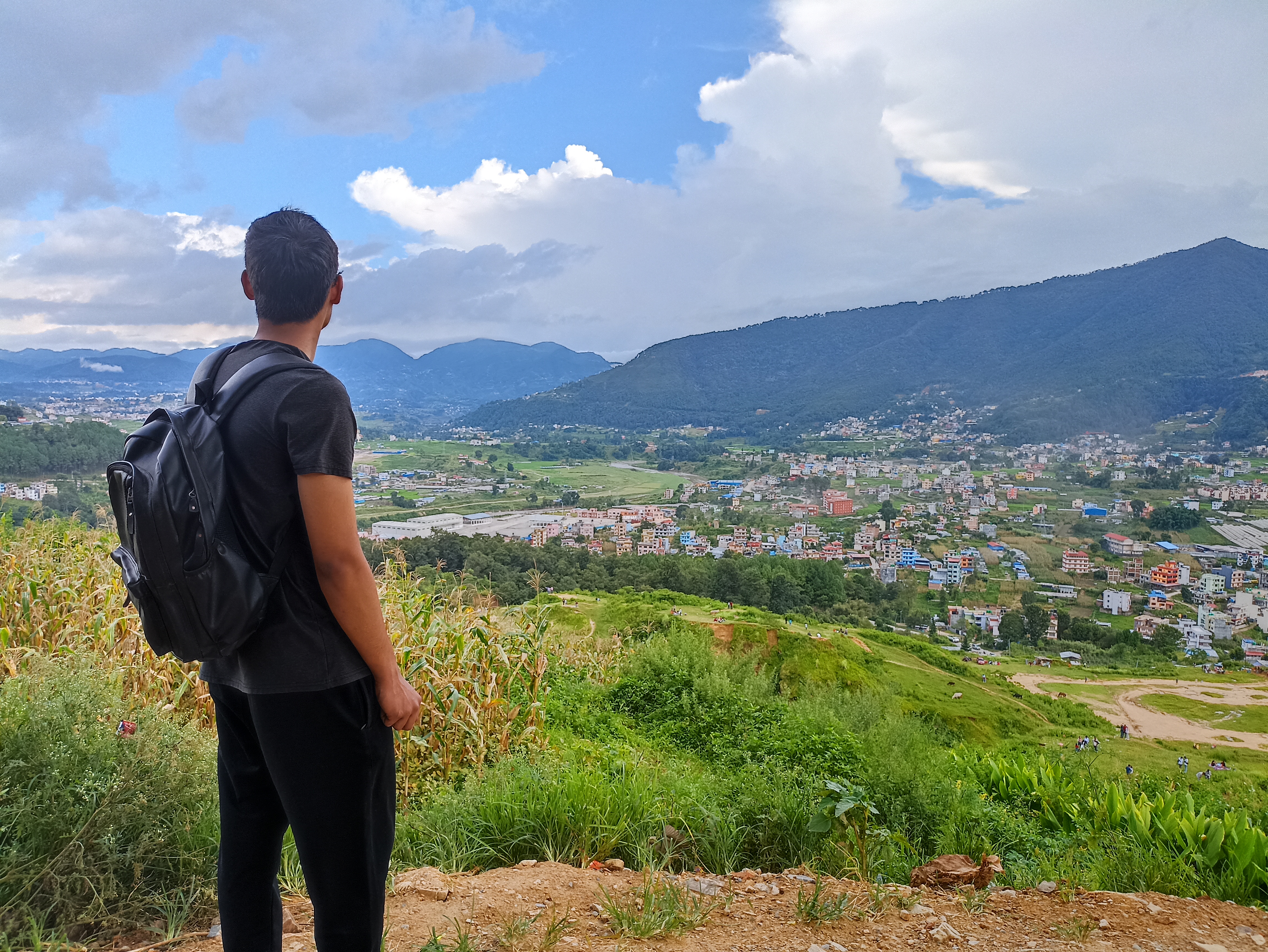 Person with a backpack standing on a hill overlooking a green valley, cityscape, and distant mountains under a partly cloudy sky.