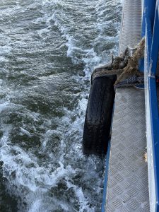 A black tyre fender hangs from the side of a blue metal boat, with foamy water churning below. 
