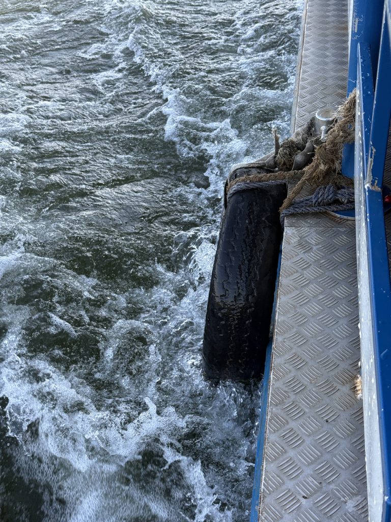 A black tyre fender hangs from the side of a blue metal boat, with foamy water churning below.