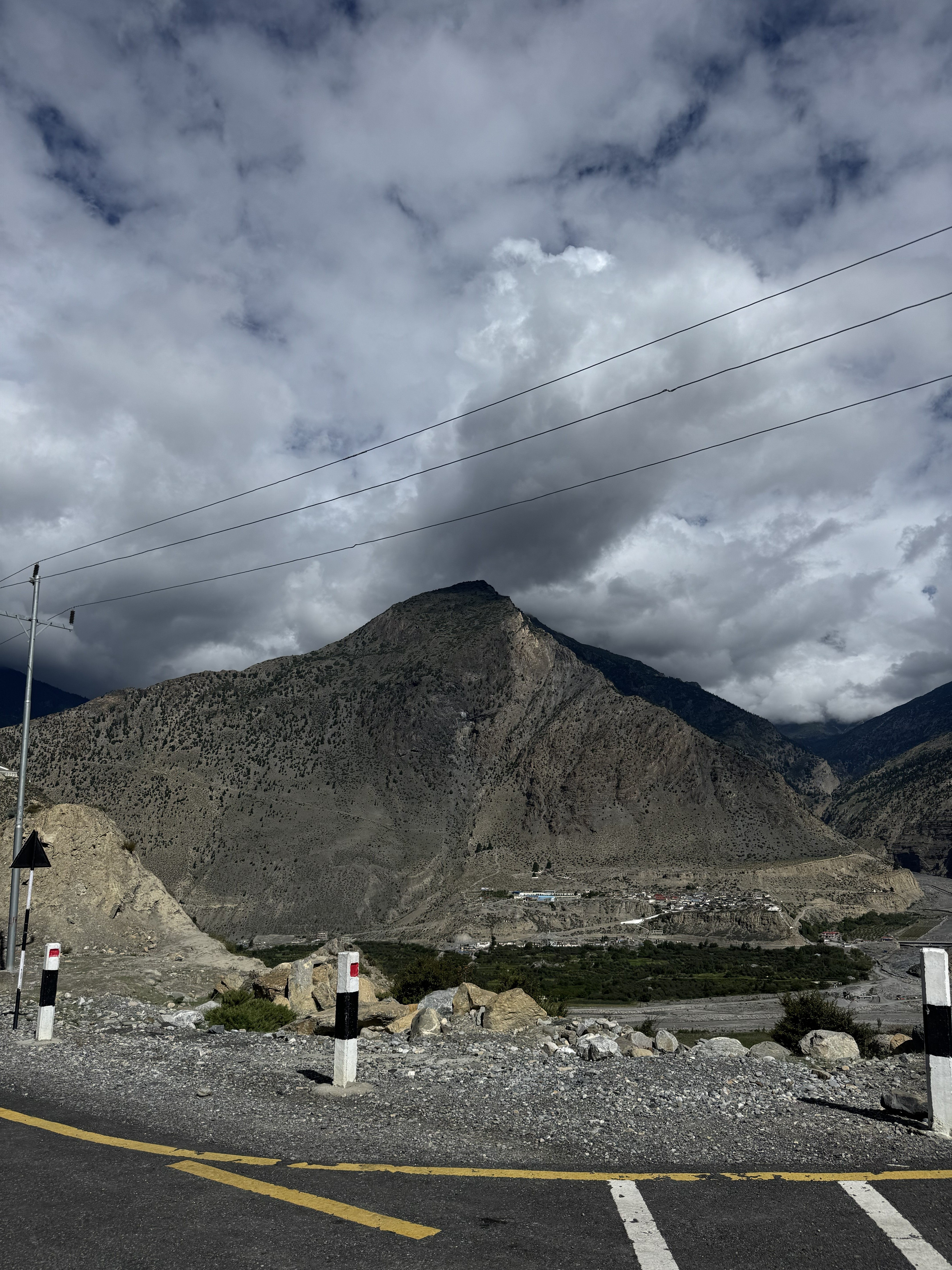 Dark mountain overlooking a remote village and green fields from a paved road under a cloudy sky.