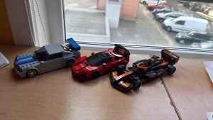 Three Lego Speed Champions cars lined up on a wooden surface by a window, with parked cars visible outside.