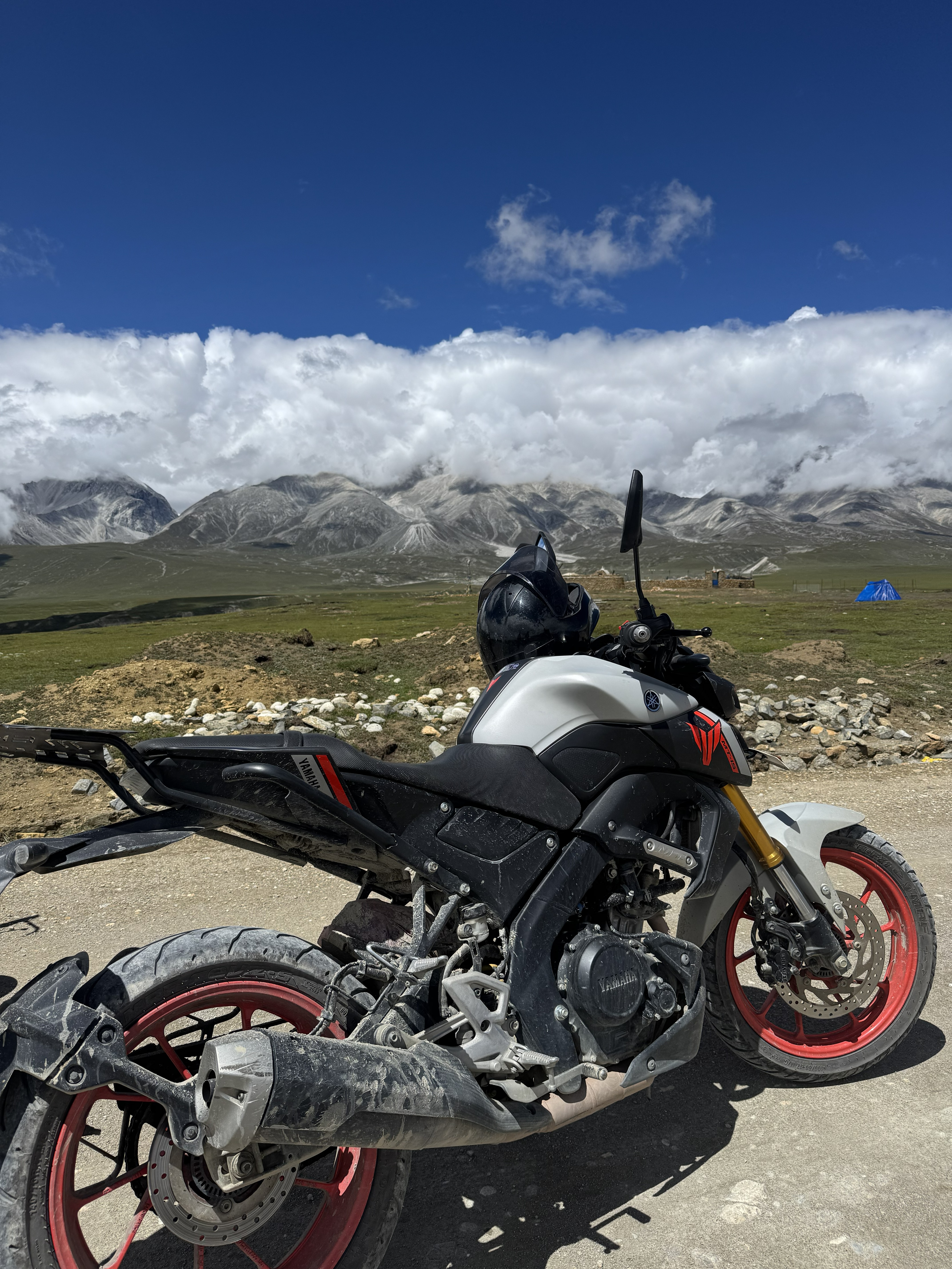 Black and silver motorcycle with a helmet on a dirt road, with green plains and rugged mountains in the background.