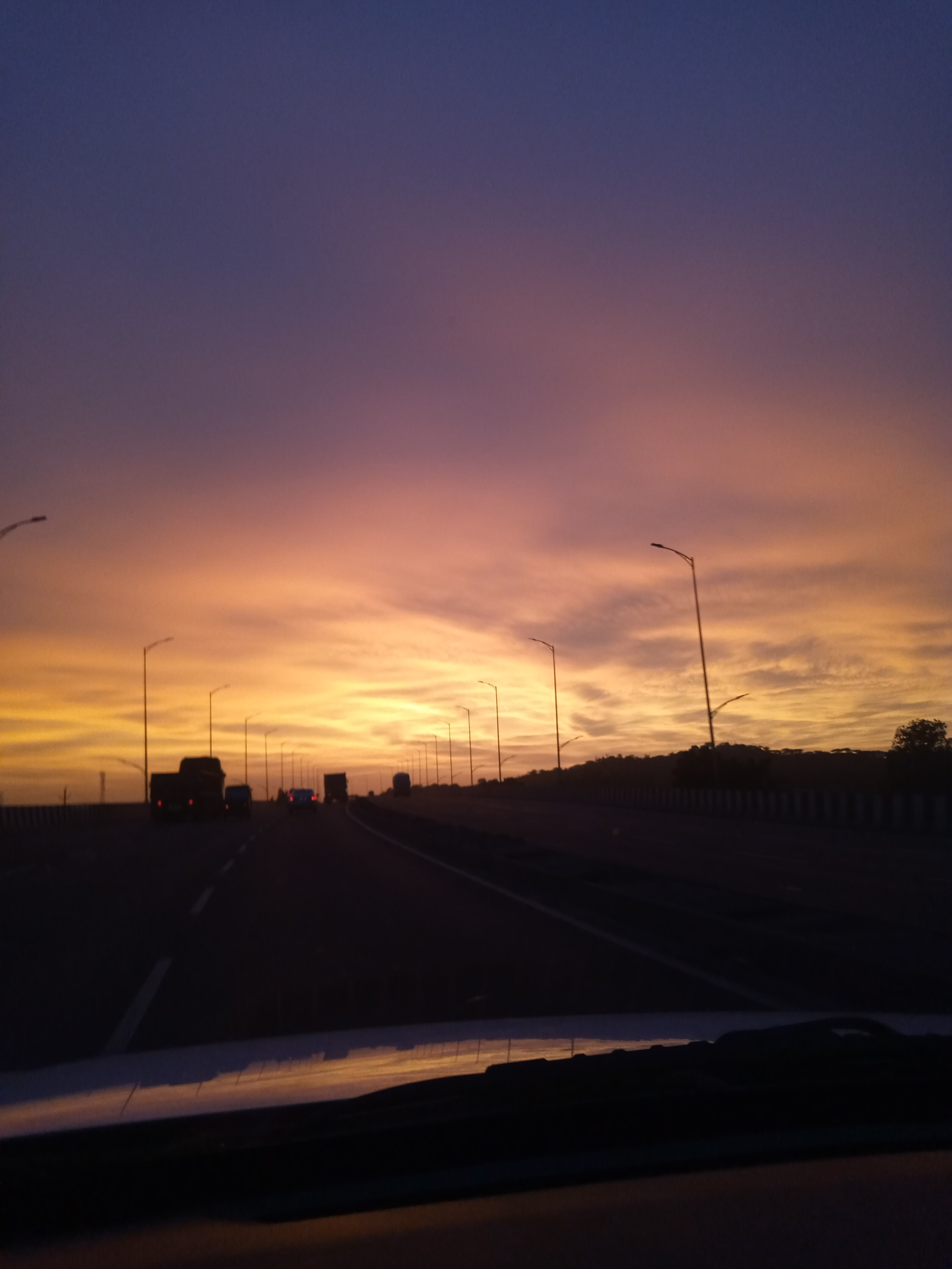 Highway at sunset with vehicles traveling beneath a blue and orange sky, lined with trees and roadside streetlights.
