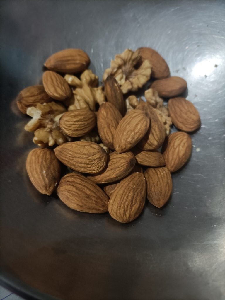 A shiny, dark bowl contains a mix of nuts. Several almonds, with oval, brown, textured shells, sit alongside a few walnut pieces. The walnut pieces are irregularly shaped and have a lighter, more crumbly appearance than the almonds' smooth surfaces.