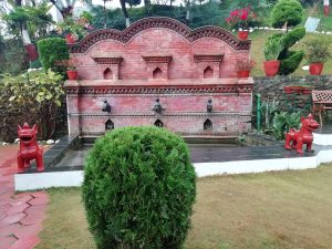 A serene outdoor scene featuring a decorative brick fountain structure with multiple water spouts, intricately carved designs, and small niches. In front of the fountain, two vibrant red sculptures resembling mythical creatures stand on either side. Surrounding the fountain, there are neatly arranged potted plants, flowers, and a well-manicured grassy area. 