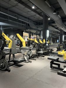 Row of black and yellow weight machines in a large gym with dark rubber flooring and industrial lighting.