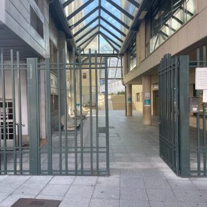 Entryway with a glass canopy, green metal gates, stone walls, benches, and a paved pathway leading toward a green area.
