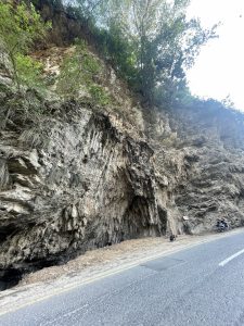 A steep, craggy rock face along the side of a paved road with a motorcycle parked further down.