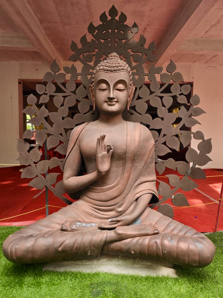 A serene Buddha statue sits in meditation, framed by an ornate decorative backdrop. The symmetrical composition and peaceful expression give the photograph a calm, contemplative feel. Location: Ayamkudy, Kottayam, Kerala.