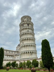 The Leaning Tower of Pisa, Italy, seen from below with its tilted tiers of arched columns, framed by cypress trees and potted plants in the foreground under a cloudy sky.
