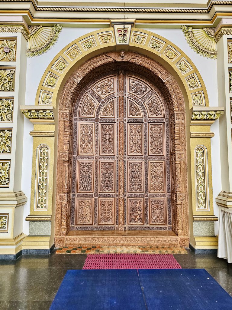 A grand carved wooden doorway surrounded by elaborate gold-and-white decorations. The intricate floral patterns and symmetrical design beautifully showcase the church's craftsmanship. Location: Ramapuram, Kottayam, Kerala.