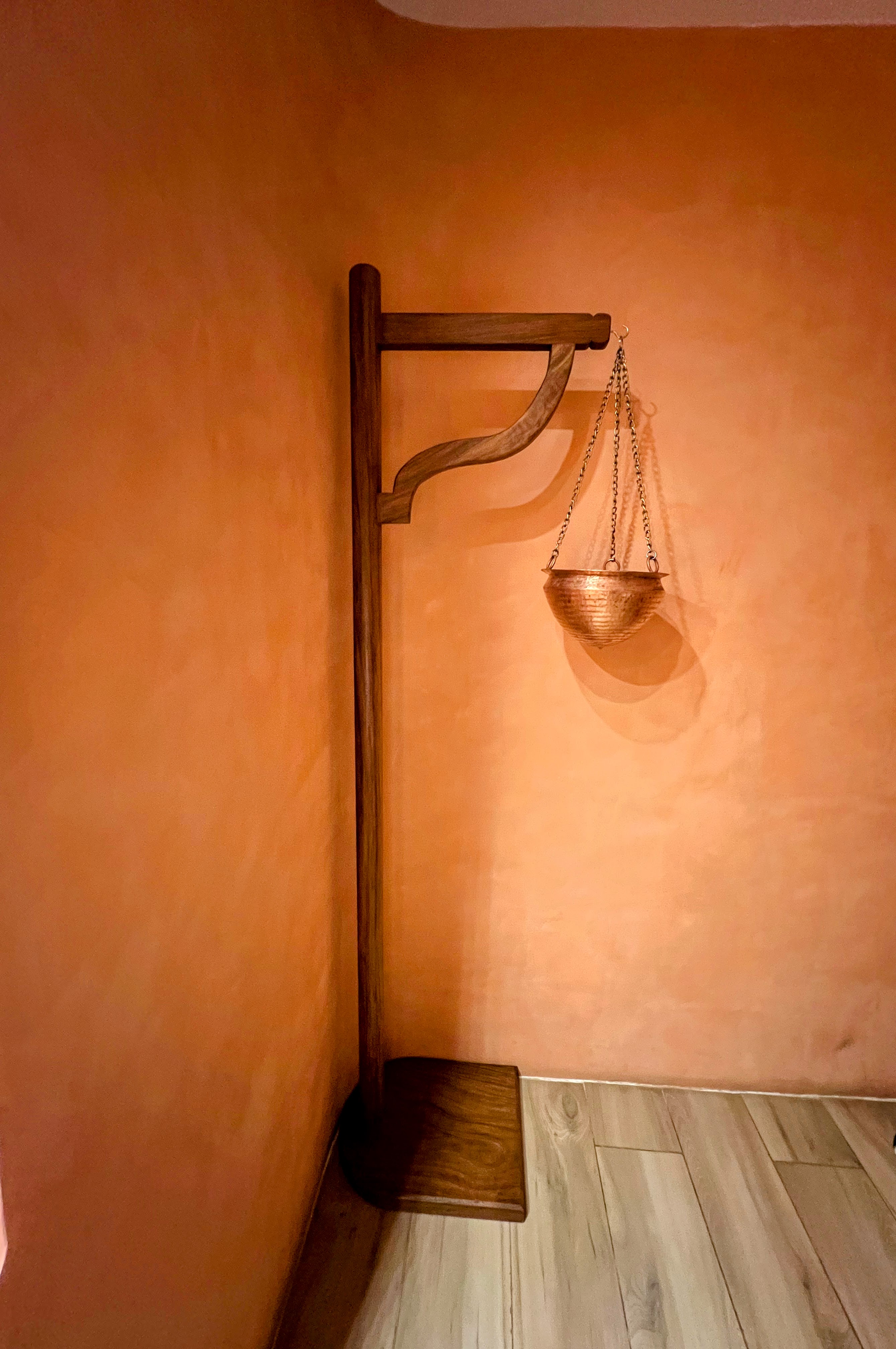 A decorative copper hanging bowl suspended by metal chains from a tall wooden stand, set against a warm terracotta wall.