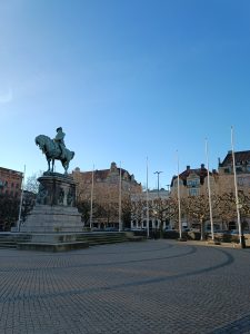 Bronze equestrian statue on a stone pedestal in a paved public square surrounded by historic European buildings and flagpoles under a blue sky.