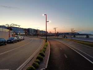 Waterfront road at twilight with parked cars, street lamps, shrubs, and buildings along the shore.