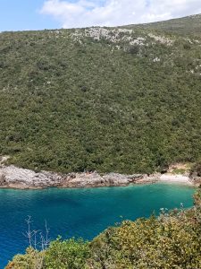 Secluded coastal cove with clear turquoise water and a small sandy beach nestled against a steep, forest-covered mountain slope.