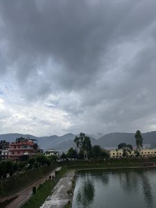 Lower-angle view of the brick-paved edge of Nag Pokhari pond, with green embankments, residential buildings, and distant hills under storm clouds.