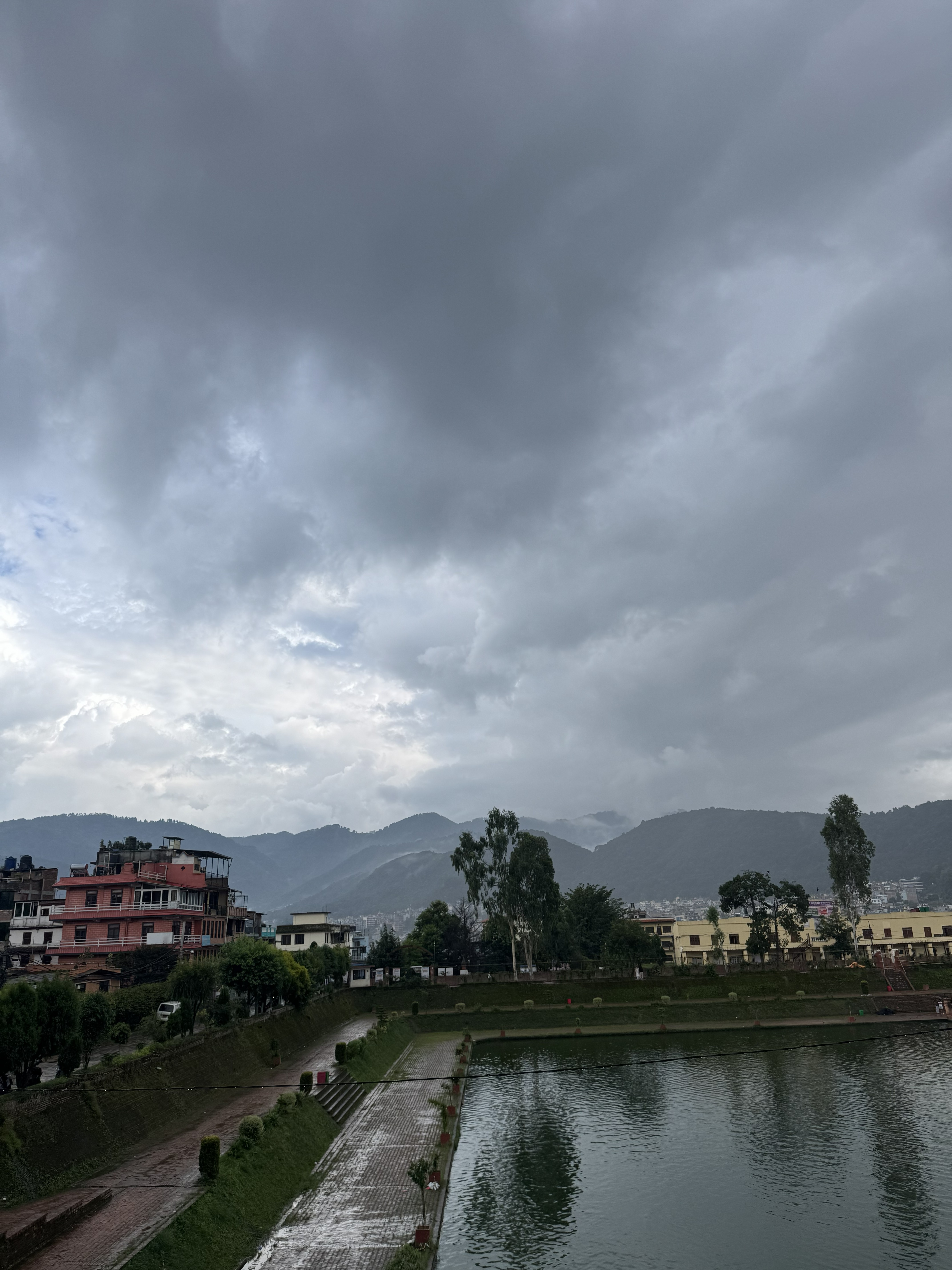 Lower-angle view of the brick-paved edge of Nag Pokhari pond, with green embankments, residential buildings, and distant hills under storm clouds.