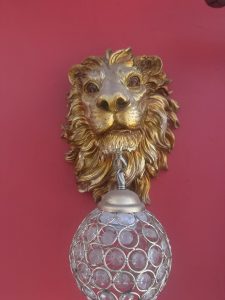 Gold lion-head wall lamp with an ornate crystal globe hanging beneath its chin on a red wall.