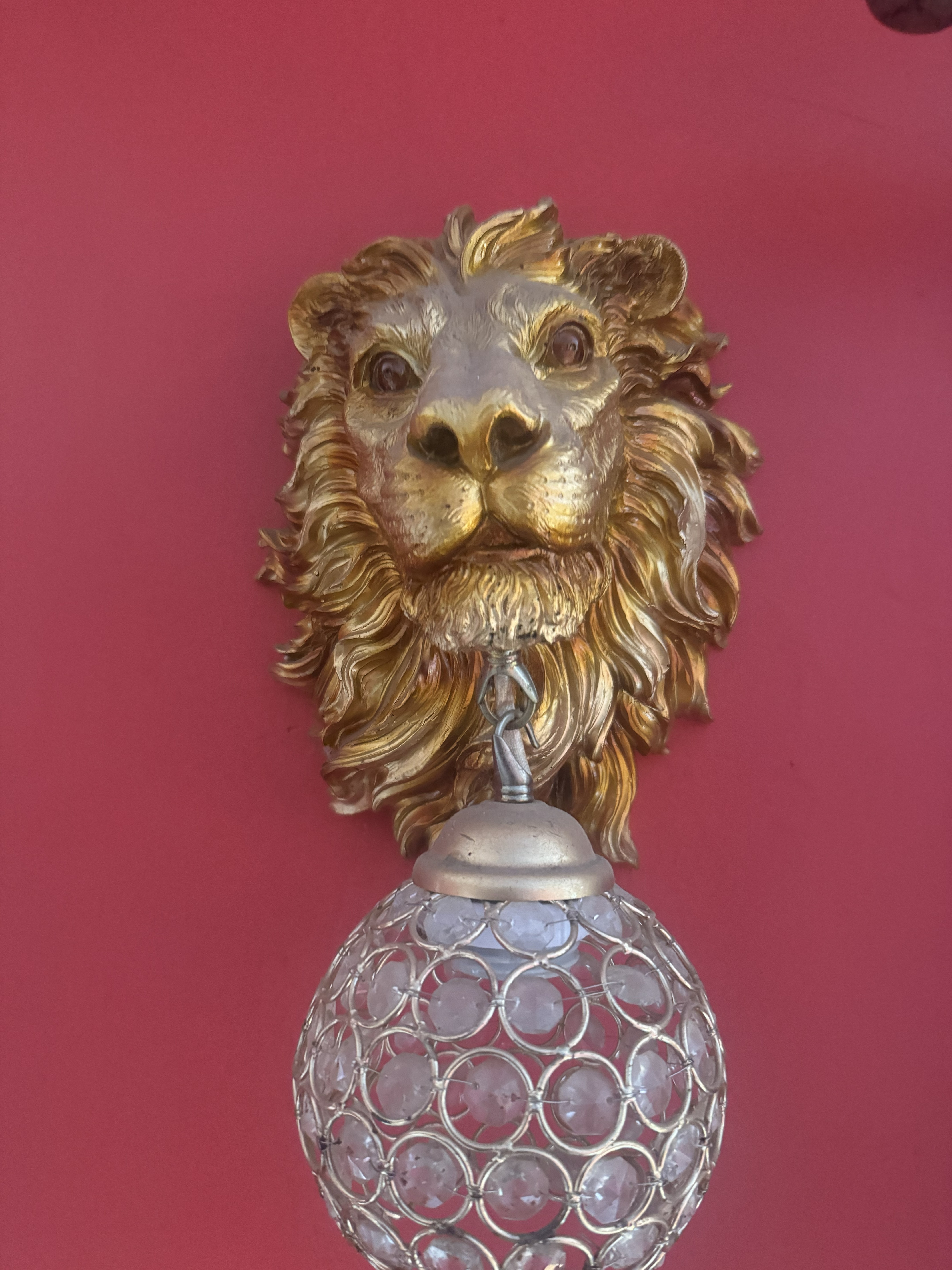 Gold lion-head wall lamp with an ornate crystal globe hanging beneath its chin on a red wall.